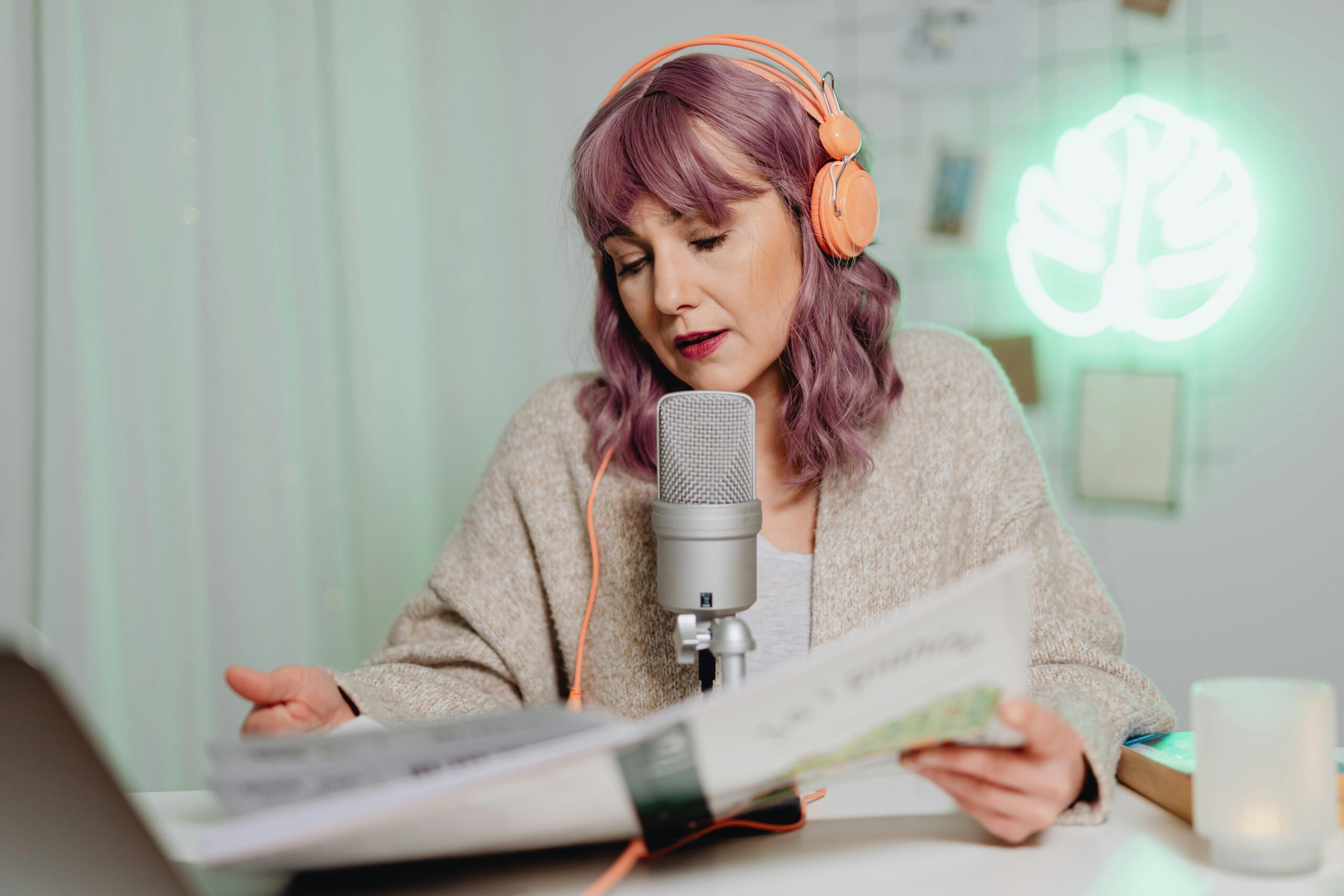 Woman with headphones podcasting indoors, speaking into a microphone while reading a book.
